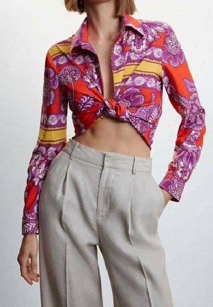 Chemise multicolore à manches longues avec des motifs floraux en violet, orange et jaune, nouée à la taille, associée à un pantalon taille haute gris clair.