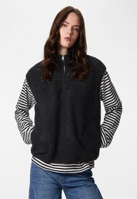 Crna prsluk od flisa s visokim ovratnikom i zatvaračem na patent, nošen preko crno-bijele prugaste majice s dugim rukavima, s bočnim džepovima.