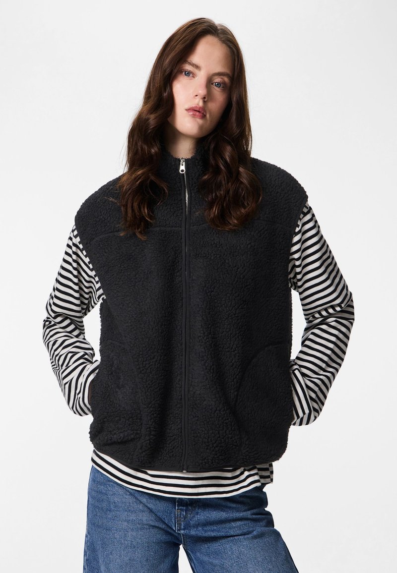 Crna prsluk od flisa s visokim ovratnikom i zatvaračem na patent, nošen preko crno-bijele prugaste majice s dugim rukavima, s bočnim džepovima.