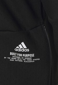 Tissu noir avec logo Adidas blanc et texte « Built for Purpose, Designed in Germany, brand with three stripes, base pattern GLX1226F20 » à côté d'une fermeture éclair.