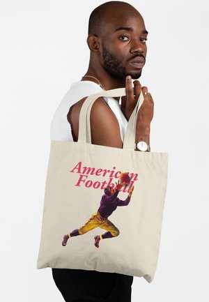 Muž s vousy drží béžovou tašku s ilustrací skákajícího hráče amerického fotbalu a červeným nápisem "American Football".