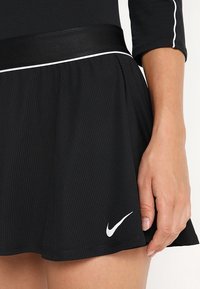 Falda atlética negra texturizada con el logo blanco de Nike cerca del dobladillo, combinada con una camiseta negra que tiene un ribete blanco en las mangas y la cintura.