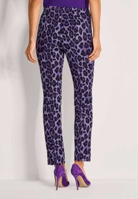 Vrouw in paarse panterprintbroek en bijpassende paarse hoge hakken, staande tegen een effen lichte achtergrond.