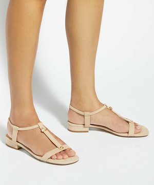 Jambes portant des sandales plates beiges avec des fines lanières et des détails de boutons dorés sur un fond blanc uni.