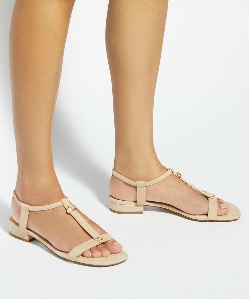 Jambes portant des sandales plates beiges avec des fines lanières et des détails de boutons dorés sur un fond blanc uni.
