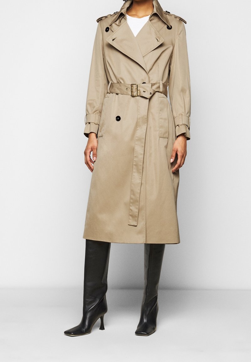 Personne portant un trench-coat beige avec une ceinture, des bottes noires montantes et un haut blanc, debout devant un fond uni.