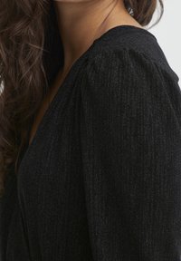 Czarna teksturowana odzież z dekoltem w serek, z bufkami i subtelnym połyskiem. Gładki, spływający materiał podkreśla nowoczesny krój.