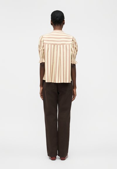Γυναίκα με τα μαλλιά της σε κότσο, που φοράει μπλούζα με κοντά μανίκια σε μπεζ και ανοιχτό καφέ κατακόρυφες ρίγες και σκούρο καφέ παντελόνι, στέκεται γυρισμένη προς τα πίσω.