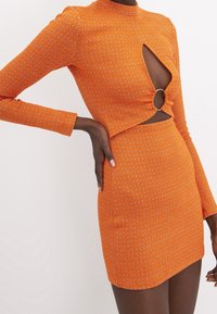 Robe en maille orange à manches longues, ornée d'une découpe à la taille et d'un accent circulaire. Motif texturé avec une silhouette ajustée.