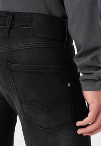 Gros plan d'une personne portant un jean gris foncé avec une poche arrière visible et une étiquette de marque en cuir noir au-dessus de la ceinture.