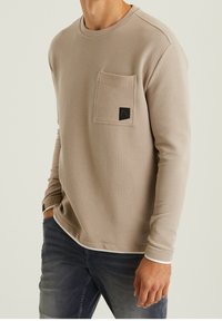 Světle béžová košile s dlouhým rukávem a texturovaným povrchem, kulatým výstřihem a kapsičkou na prsou se tmavou nálepkou. Spárováno s šedými džíny.