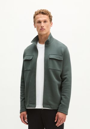 Man die een groene ritsjacket met klepzakken op de borst over een wit overhemd draagt, staand tegen een effen lichte achtergrond.