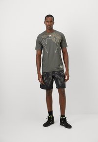 T-shirt de sport vert foncé avec un design géométrique doré, assorti à un short noir avec un motif graphique, porté avec des chaussures noires.