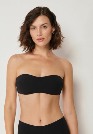 Mujer con cabello castaño hasta los hombros, vistiendo un top bandeau negro sin tirantes, sobre un fondo liso y claro.