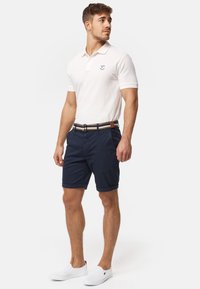 Weißes Poloshirt mit kurzen Ärmeln, marineblaue Shorts mit umgeschlagenem Saum und weiße Slipper. Brauer und gestreifter Gürtel als Akzentdetail.