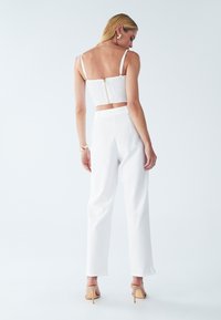 Biała, marszczona bluzka crop top z zamkiem błyskawicznym z tyłu, w połączeniu z białymi spodniami o wysokim stanie. Modelka ma na sobie cieliste szpilki i złotą bransoletkę.