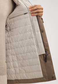 Gepolsterte Jacke mit einem beigen, gesteppten Innenfutter und einer Reißverschlusstasche. Die Außenseite besteht aus einem einfarbigen braunen Stoff mit glatter Oberfläche und Reißverschluss.