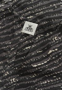 Schwarz- und graumusterter Stoff mit gewellten Streifen, der eine strukturierte Oberfläche aufweist, sowie einem weißen Etikett mit einem stilisierten schwarzen Logo.