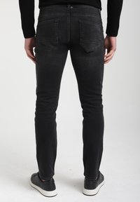Mustad denim püksid, millel on slim fit, varustatud tagakottide ja minimaalsete plekkidega. Mudel kannab musti kingi, näidates pükse tagant.