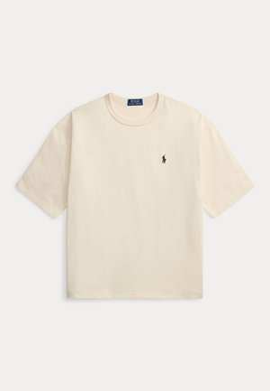 Jeune homme portant un t-shirt crème, un jean large bleu foncé, des chaussures blanches, tenant une veste verte sur son épaule sur un fond neutre.