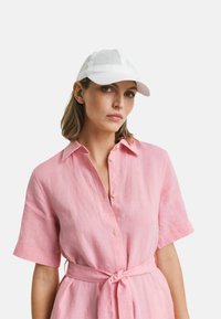 Camicia button-up in lino rosa con maniche corte e dettaglio con cintura. Berretto da baseball bianco completa il look. Tessuto morbido, vestibilità rilassata.