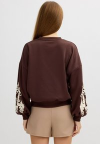 Sudadera marrón con mangas voluminosas que presenta patrones florales bordados en blanco; combinada con pantalones cortos beige. Textura suave y ajuste relajado.