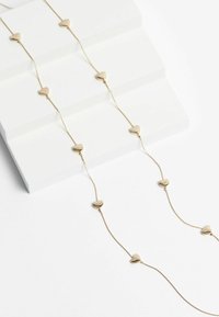 Goldfarbene Halskette mit kleinen Herz-Anhängern, die gleichmäßig an einer filigranen Kette verteilt sind, auf einer weißen Oberfläche präsentiert.