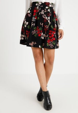 Jupe plissée noire avec imprimé floral rouge, blanc et rose, portée avec un haut blanc à manches longues et des bottines noires.