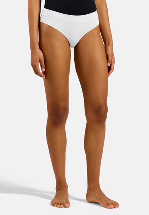 Vrouw die witte hoog uitgesneden slips en een zwarte top draagt, blootsvoets staand op een effen lichte achtergrond.