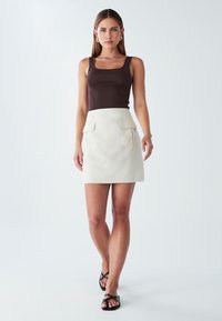 Rjav rebrast top brez rokavov v kombinaciji s krema mini krilom, ki ima dve sprednji žepki, ter črnimi sandali na vezalko. Gladke teksture tkanin v celoti.