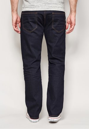 Donkerblauwe spijkerbroek met een rechte pijp, zichtbare stiksels en twee achterzakken, gedragen met witte sneakers.