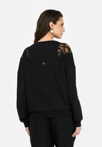 Sweatshirt noir avec des détails en dentelle aux épaules, coupe ample, col rond et accent en logo doré. Manches longues et poignets côtelés.