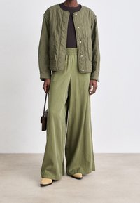 Osoba w oliwkowej, pikowanej kurtce, szerokich zielonych spodniach, beżowych czółenkach, trzymająca małą, ciemnobrązową torebkę na jednolitym tle.