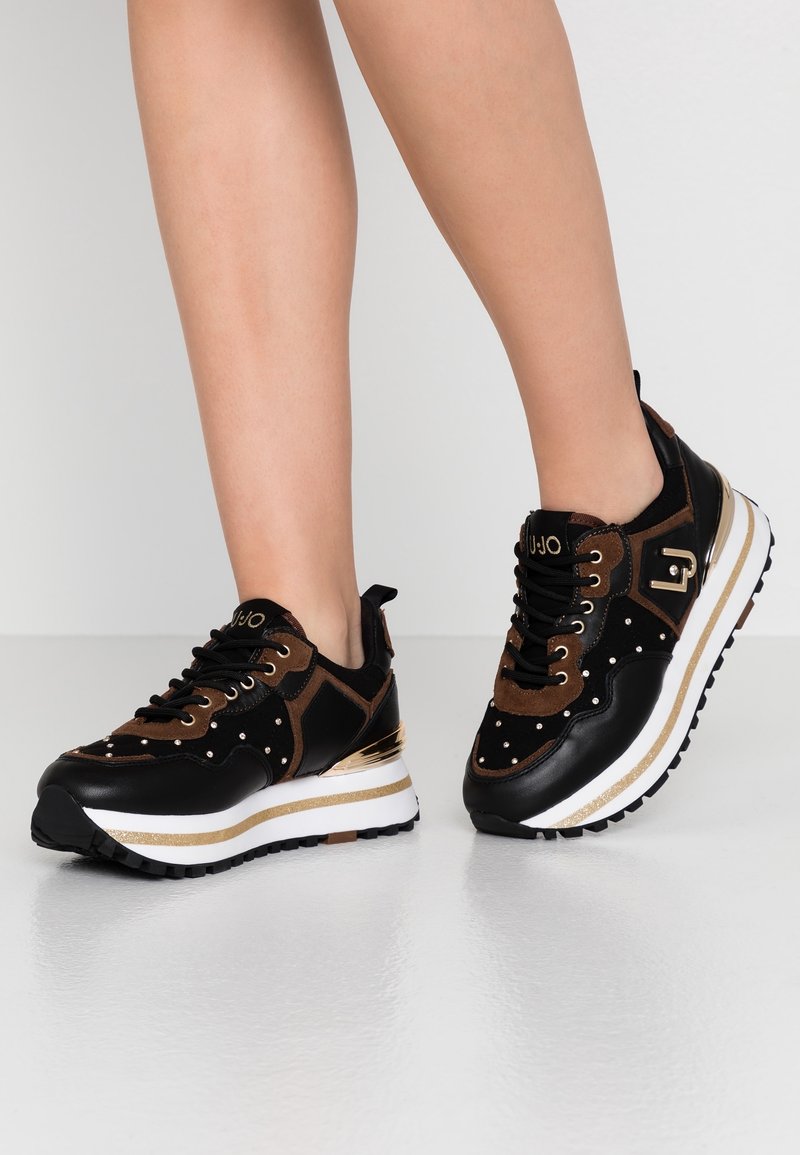 Sneaker neri e marroni con una combinazione di texture lisce e scamosciate, accenti dorati e dettagli borchiati su una suola spessa bianca e dorata.