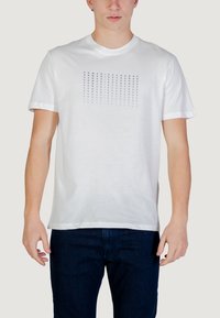 Homme portant un t-shirt blanc à manches courtes avec le texte répété « ARMANI EXCHANGE » en bleu dégradé, associé à un jean bleu foncé, debout devant un fond blanc.