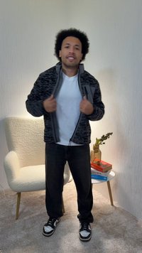 Μαύρο και γκρι μπουφάν με σχέδιο, φερμουάρ μπροστά, φορεμένο πάνω από λευκό t-shirt. Καθημερινό μαύρο παντελόνι και μαύρα/ασημί αθλητικά παπούτσια συμπληρώνουν την εμφάνιση.