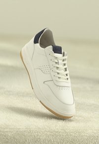 White leather sneaker with beige sole and black heel tab, featuring laces and branded tongue, displayed on soft beige textured surface.