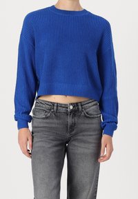 Maglione corto blu lavorato a maglia con texture a coste, scollatura rotonda e maniche lunghe, abbinato a jeans in denim grigi.