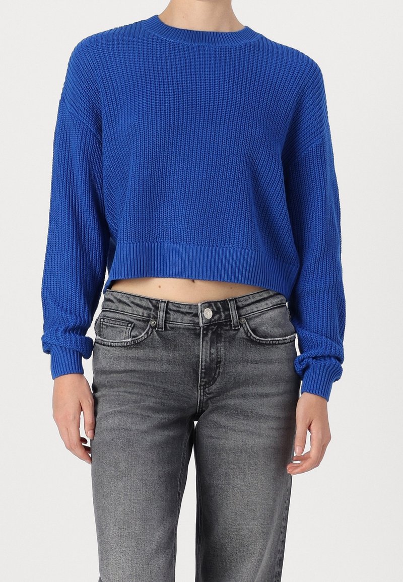 Maglione corto blu lavorato a maglia con texture a coste, scollatura rotonda e maniche lunghe, abbinato a jeans in denim grigi.