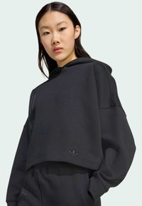 Hoodie corto nero realizzato in tessuto morbido, dotato di cappuccio, polsini a coste e un logo discreto sull'orlo. Vestibilità rilassata con una texture morbida.