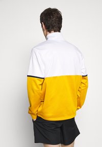 Żółta i biała kurtka na zamek błyskawiczny z wysokim kołnierzem, z granatowym paskiem akcentującym. Miękka tkanina o sportowym, kolorowym wzornictwie.