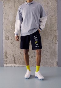 Osoba v světle šedém tričku s dlouhým rukávem, černých šortkách s několika logy Nike, žlutých ponožkách a bílých teniskách stojící uvnitř.