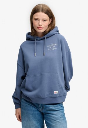 Ung kvinde iført en blå Superdry hættetrøje med lomme foran og blå jeans, stående foran en ensfarvet hvid baggrund.