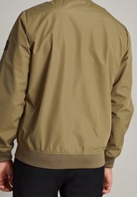 Chaqueta bomber ligera en color caqui con una textura suave, puños elásticos y cinturilla. Sin hardware visible ni patrones. Vista trasera.