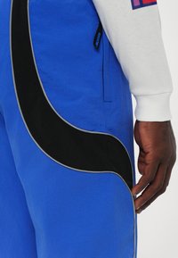 Blå joggebukser med et svart buet design og grå piping. Inkluderer sidelommer og et teksturert materiale.