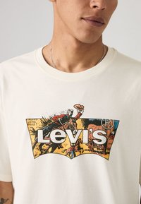T-shirt en coton couleur crème avec une impression graphique "Levi's" en gras, accompagnée d'une illustration vintage de cowboys.