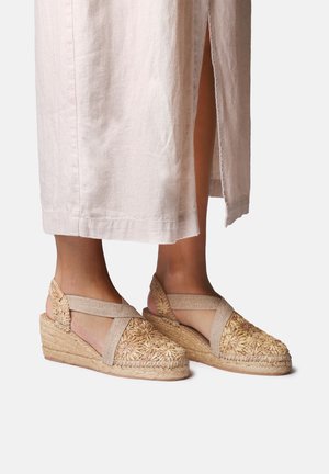 Įstrižainės espadrilės su pintu šiaudiniu dekoru ir elastinėmis čiurnos juostomis. Smėlio spalvos, tekstūruota medžiaga su gėlių raštais ant pirštų dalies.
