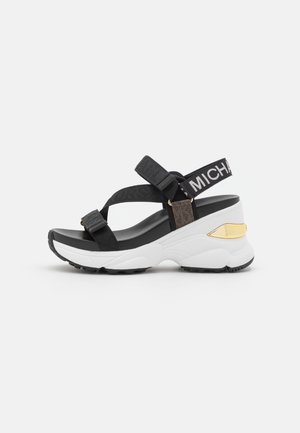 Sandale plateforme noir et blanc avec des sangles ajustables, présentant un accent doré sur le talon et un textile texturé. Semelle en caoutchouc pour une bonne adhérence.