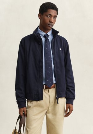 Jonge man met een marineblauwe jas, lichtblauw shirt, marineblauwe stropdas met witte stippen, beige broek, die een riem van een bruine leren tas vasthoudt in een neutrale omgeving.