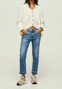 Mujer con blazer y chaleco color crema, vaqueros azules recortados con dobladillos deshilachados, y zapatillas rosas y grises, de pie con las manos en los bolsillos.